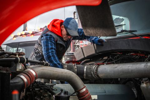 Technician in a cap and gloves leaning under the raised hood of a red semi tractor, checking the engine